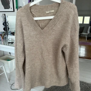 Beige stickad tröja med v-ringning från Gina Tricot - Mysig beige stickad tröja från Gina Tricot med djup v-ringning och ribbade muddar. Perfekt för lager-på-lager och passar till både jeans och kjol. Enkel och stilren design som funkar året runt.