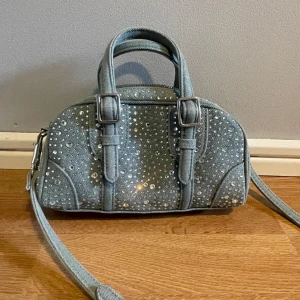 Jeansväska med strass och axelrem - Cool handväska i ljusblått jeansmaterial med massor av glittrande strassdetaljer. Väskan har både handtag och avtagbar axelrem samt dragkedja upptill. Perfekt för dig som vill sticka ut med en unik accessoar.