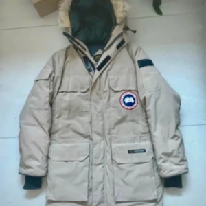 Beige dunjacka från Canada Goose - Säljer en beige dunjacka från Canada Goose med huva och pälskant. Jackan har flera stora fickor framtill, svart innerfoder och klassisk logga på bröstet. Perfekt för kalla dagar och riktigt varm.