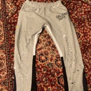 Grå mjukisbyxor från Gallery Dept. - Grå mjukisbyxor från Gallery Dept. med svart tryck på höften och vita färgstänk längs benen. Byxorna har elastisk midja och svarta paneler längs sidorna. Perfekta för en avslappnad och trendig stil. Den är helt ny och oanvänd. 