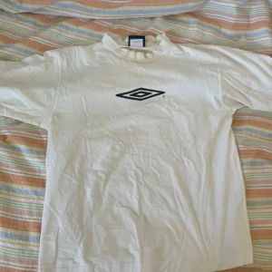 Vit t-shirt från Umbro med logga - Säljer en vit t-shirt från Umbro med klassisk svart Umbro-logga på bröstet och en mindre logga på ärmen. T-shirten har normal passform och är tillverkad i mjuk bomull. Perfekt för en sportig look.