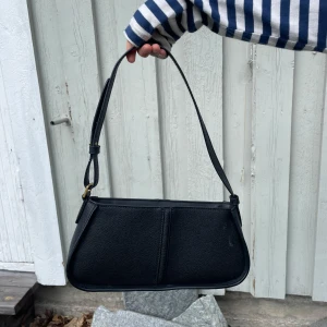 Svart axelväska i skinn - Snygg svart axelväska i skinn med enkel och stilren design. Väskan har gulddetaljer och en mjukt rundad form. Perfekt för att bära det viktigaste och passar till många olika outfits.    Ser nu att det är en liten fläck på väskan men tar bort den innan jag skickar såklart!!💘