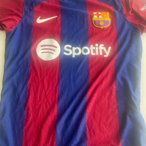 FC Barcelona hemmatröja 2022/23 - Säljer en officiell FC Barcelona hemmatröja från säsongen 2022/23 med Spotify som sponsor. Tröjan är kortärmad, har klassiska blå och röda vertikala ränder, klubbmärke på bröstet och La Liga-märke på ärmen. Perfekt för alla Barca-fans!