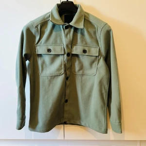 Grön overshirt från 157 - Snygg grön overshirt från 157 med två stora bröstfickor och knappar framtill. Perfekt att bära som lager över en t-shirt. Klassisk krage och lång ärm för en stilren look.