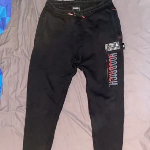 Svarta mjukisbyxor från Hoodrich - Snygga svarta mjukisbyxor från Hoodrich med tryck i vitt och rött. Byxorna har en elastisk midja med snörning och flera detaljerade tryck på benen. Perfekta för en avslappnad stil.
