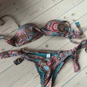 Färgglad mönstrad bikini stl S-M ( nt använd ) - Snygg bikini med ett färgglatt och detaljerat mönster i turkos, orange och rosa. Överdelen har en bandeau-stil med avtagbara axelband och knytning i ryggen. Underdelen har knytband i sidorna för justerbar passform.