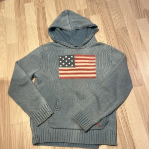 Blå stickad hoodie från Polo Ralph Lauren - Säljer en snygg blå stickad hoodie från Polo Ralph Lauren med en stor amerikansk flagga på framsidan. Tröjan har långa ärmar och en praktisk magficka. Perfekt för en avslappnad stil! 💙