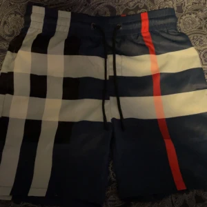 Randiga badshorts från Burberry - Snygga badshorts från Burberry med ett klassiskt randigt mönster i marinblå, vitt och rött. Shortsen har en bekväm passform med snörning i midjan för justerbarhet. Perfekta för sommaren. 