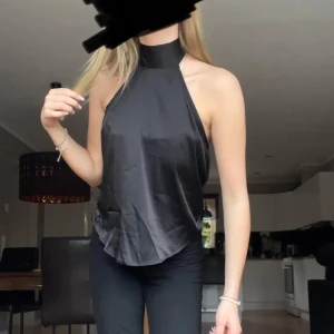 Svart halterneck-top - Snygg svart halterneck-top i ett glansigt material. Toppen har en öppen rygg och knyts i nacken, vilket ger en elegant look. Aldrig använd, perfekt för fest!