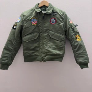 Grön bomberjacka med patches - Säljer en grön bomberjacka med flera coola patches, inklusive 'Top Gun' och amerikanska flaggan. Jackan har dragkedja framtill och fickor med lock. Perfekt för en avslappnad och stilren look.
