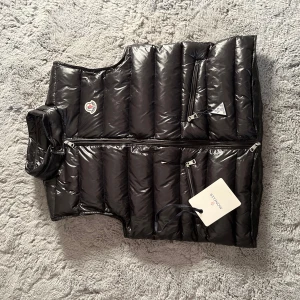 Moncler Gui Väst - Snygg svart dunväst från Moncler med glansig finish. Västen har dragkedja och flera praktiska fickor med dragkedjor. Perfekt för kyligare dagar när du vill hålla stilen.