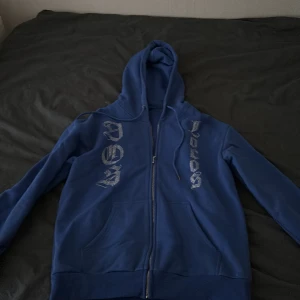 Dos locos, Blå hoodie med glittriga detaljer - Säljer en blå hoodie med dragkedja och glittriga detaljer på både fram- och baksidan. Den har en stor huva och långa ärmar, märket dos locos som nu längre inte finns, denna släpptes i en upplaga där bara 100 gjordes. Pris kan diskuteras 