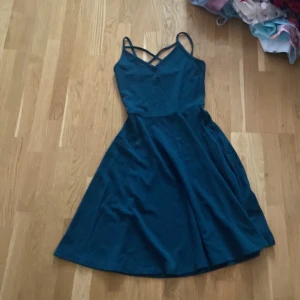 Mörkgrön klänning från Gina Tricot - Snygg mörkgrön klänning från Gina Tricot med smala axelband och korsdetalj framtill. Perfekt för en stilren look. Klänningen har en v-ringad design och är knälång, vilket ger en elegant siluett.