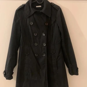 Svart trenchcoat från Vila - Snygg svart trenchcoat från Vila med dubbelknäppning och klassisk krage. Jackan har långa ärmar och justerbara remmar vid ärmsluten. Perfekt för höst och vår!