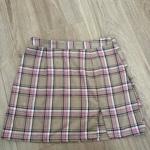 Beige och rosa rutig kjol - Snygg beige kjol med rosa och svarta rutor. Kjolen har en liten slits framtill och en dragkedja i sidan för enkel påklädning. Perfekt för en stilren look!