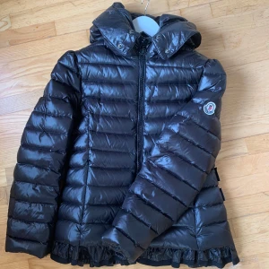 Svart dunjacka från Moncler - Snygg svart dunjacka från Moncler med dragkedja och huva. Jackan har en quiltad design med en elegant passform.. Perfekt som vår eller höstjacka med  kyliga dagar när du vill hålla stilen. OBS volangerna längst ner är avklippta. 