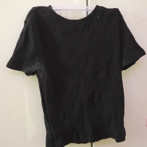 Svart ribbad t-shirt - Säljer en stilren svart ribbad t-shirt med korta ärmar. Perfekt för en avslappnad look. Passar till det mesta och är ett måste i garderoben!