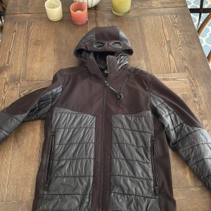 Svart jacka med huva och glasögon från C.P. Company - Säljer en cool svart jacka från C.P. Company med unik huva som har inbyggda glasögon. Jackan har dragkedja och man kan även ta bort huvan om man känner för det.  Perfekt för hösten.