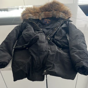 Svart parkajacka med päls från Woolrich - Säljer en svart parkajacka från Woolrich med en lyxig pälsfodrad huva. Jackan har dragkedja och knappar framtill samt flera praktiska fickor. Perfekt för kalla vinterdagar.