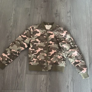 Camouflage bomberjacka från KappAhl - Säljer en cool bomberjacka från KappAhl med ett rosa och grönt camouflagemönster. Jackan har dragkedja framtill och en ficka med dragkedja på ärmen. Perfekt för att ge en edgy touch till din outfit!