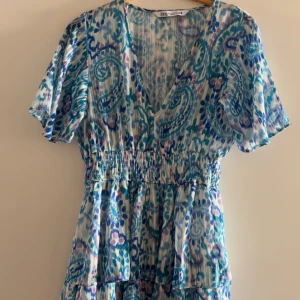 Mönstrad klänning från Zara - Säljer en färgglad klänning från Zara med ett blått och grönt mönster. Klänningen har korta ärmar och en v-ringad design. Den är perfekt för sommarens varma dagar.