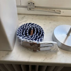 Blåvitt flätat bälte - Säljer ett snyggt blåvitt flätat bälte med bruna läderdetaljer. Perfekt för att ge din outfit en cool touch. Bältet har en klassisk design med en silverfärgad spänne.