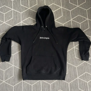 Svart hoodie från Palm Angels - Snygg svart hoodie från Palm Angels med stor logga på ryggen och mindre logga på framsidan. Tröjan har en klassisk känguruficka och justerbar huva med dragsko. Perfekt för en stilren och avslappnad look.