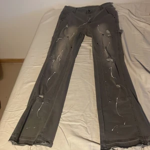 Grå flared jeans med slitningar - Säljer ett par grå utsvängda (flared) jeans med slitna detaljer och färgstänk. Byxorna har flera fickor och en avslappnad passform. Perfekta för en cool och avslappnad stil.