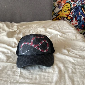 Svart keps med ormmönster - Snygg svart keps med ett coolt ormmönster i rött och vitt. Kepsen har ett nätmaterial på sidorna och ett justerbart spänne bak för perfekt passform. Perfekt för att ge din outfit en edgy touch. Varan ör köpt på denna app plick så är inte säker att den är äkta
