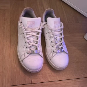 Vita/rosa  Adidas Stan Smith sneakers - Säljer ett par klassiska vita Adidas Stan Smith sneakers med rosa detaljer på hälen. Skorna är i hyffsat bra skick, men det syns att någon har andvändigt de! Och de är såklart finare på! Säljer för 450 men kan också tänka mig 400