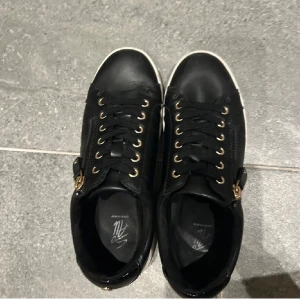 Svarta sneakers med dragkedja - Snygga svarta sneakers med snörning och dragkedja på sidan. Skorna har en vit sula och guldiga detaljer vid snörningen. Som ny har inte haft på mig de så många tillfällen 
