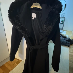 Svart kappa med pälsdetaljer - Elegant svart kappa från H&M med en lyxig pälsdetalj runt huvan. Kappan har långa ärmar och ett knytbälte i midjan. 