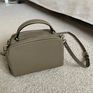 Brun axelväska med dragkedja - Snygg beige axelväska i syntetmaterial med praktisk dragkedja och justerbar axelrem. Perfekt för att bära dina nödvändigheter med stil. Väskan har även ett handtag för extra bäralternativ.