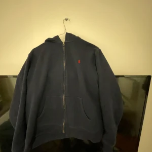 Mörkblå hoodie från Ralph Lauren - Säljer en snygg mörkblå hoodie från Ralph Lauren med dragkedja och det klassiska röda logotypen på bröstet. Skön nu för våren. Liten i storleken då den är Barn XL men sitter som S. Passar mig som är 173
