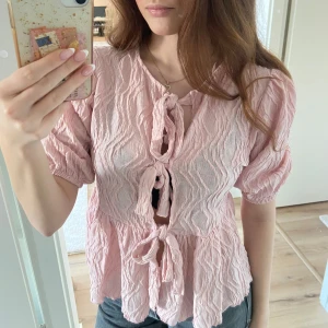 Rosa blus med knytning - Säljer en söt rosa blus med knytning framtill. Blusen har korta ärmar och en volangdetalj vid nederkanten. Perfekt för en somrig look!