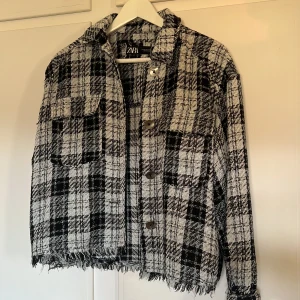 Rutig jacka från Zara - Snygg rutig jacka från Zara i svart och vit och lite blått. Jackan har en klassisk krage och knappar framtill. Den är croppad med fransiga kanter och har två bröstfickor. Perfekt för en trendig look!