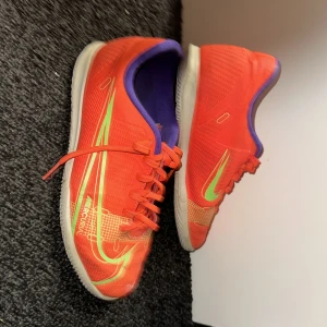 Orange Nike Mercurial fotbollsskor - Snygga orangea Nike Mercurial fotbollsskor med gröna detaljer och snörning. Skorna har en sportig design med en vit sula och lila insida, perfekt för inomhusfotboll.