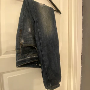 Blå jeans med slitningar - Säljer ett par blå jeans med coola slitningar och en klassisk femficksdesign. De har en knappgylf och är perfekta för en avslappnad stil. Passar bra till sneakers eller kängor.