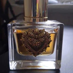 Parfym från Dolce & Gabbana - Elegant parfymflaska från Dolce & Gabbana med en gyllene dekorativ detalj i form av ett hjärta på framsidan. Flaskan är rektangulär och genomskinlig med en gyllene kork. Perfekt för den som älskar lyxiga och stilrena dofter. 30ml, har andvänts få gånger, kommer utan förpackning