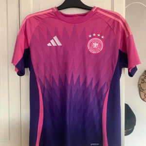 Tysklands landslag tröja - Säljer en snygg tysk fotbollströja i rosa och lila med korta ärmar. Tröjan har ett geometriskt mönster och ett emblem på bröstet. Perfekt för fotbollsfans som vill fräscha upp sig till sommaren. S sitter som M. Pris ej hugget i sten!