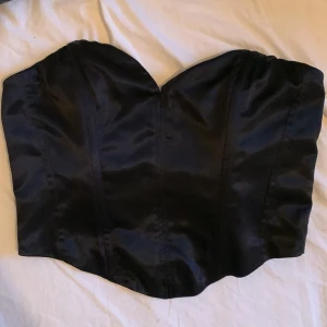 Svart korsett från H&M Divided - Snygg svart korsett från H&M Divided med dragkedja bak. Perfekt för en stilren look. Tillverkad i ett glansigt material som ger en lyxig känsla.