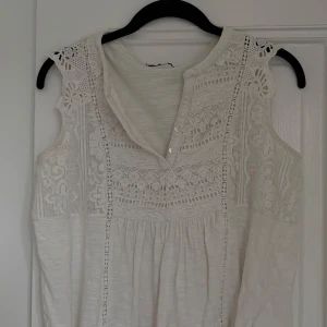 Vit ärmlös blus med spetsdetaljer - En vacker vit ärmlös blus med fina spetsdetaljer och knappar framtill. 