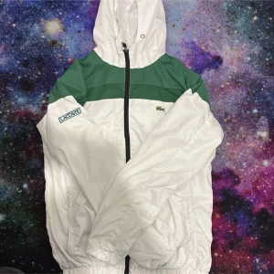 Vit och grön vindjacka från Lacoste - Säljer en stilren vit och grön vindjacka från Lacoste. Jackan har en dragkedja framtill och en justerbar huva. Perfekt för blåsiga dagar med sin lätta och bekväma design. Lacoste-loggan syns på bröstet och ärmen.