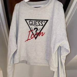 Grå sweatshirt från Guess - Säljer en grå sweatshirt från Guess med deras ikoniska logga i svart och rött framtill. Tröjan har långa ärmar och en rund halsringning. 