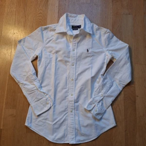 Vit skjorta från Polo Ralph Lauren - Säljer en klassisk vit skjorta från Polo Ralph Lauren i storlek XS. Skjortan är tillverkad i 100% bomull och har långa ärmar. Den har knappar framtill och en broderad logotyp på bröstet. Perfekt för en stilren look!