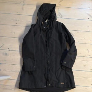 Svart regnjacka från Tretorn - Säljer en stilren svart regnjacka från Tretorn. Jackan har en praktisk huva och stängs med dragkedja och knappar. Perfekt för regniga dagar med sin långa design och justerbara ärmslut.