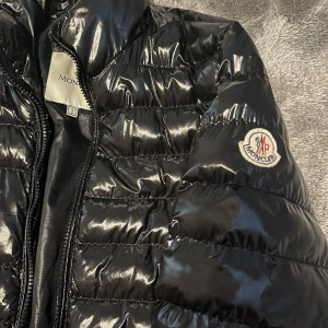 Svart pufferjacka från Moncler - Säljer en snygg svart pufferjacka från Moncler med glansig finish. Jackan har en dragkedja framtill och Moncler-logga på ärmen. Perfekt för kyligare dagar med sin quiltade design.