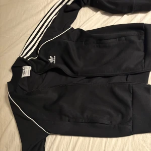 Svart jacka från Adidas - Säljer en svart jacka från Adidas med de klassiska tre vita ränderna längs ärmarna. Jackan har dragkedja framtill och är långärmad. Perfekt för en sportig look!