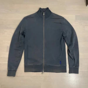 Grå jacka från Tiger of Sweden - Snygg grå zip up från Tiger of Sweden med dragkedja framtill. Jackan har en stilren design med ribbade muddar vid ärmslut och nederkant. Perfekt för en avslappnad och stilren look. Använd väldigt lite, hör av er vid funderingar