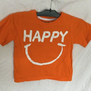 Orange t-shirt från Rebel by Primark - Säljer en glad orange t-shirt från Rebel by Primark med texten 'HAPPY' och ett leende tryckt i vitt på framsidan. Perfekt för att sprida glädje! 👕T-shirt i bomull, 92 cm Tvättråd fattas! Längd: 35 cm